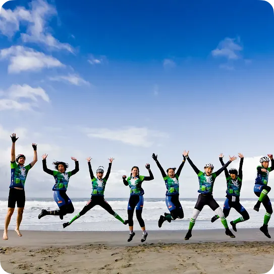 A group of eight cyclists in matching green, blue, and black athletic jerseys and helmets jumping excitedly in the air on a sandy beach. They are lined up side-by-side with arms raised in celebration, set against a background of ocean waves and a bright blue sky with scattered white clouds.