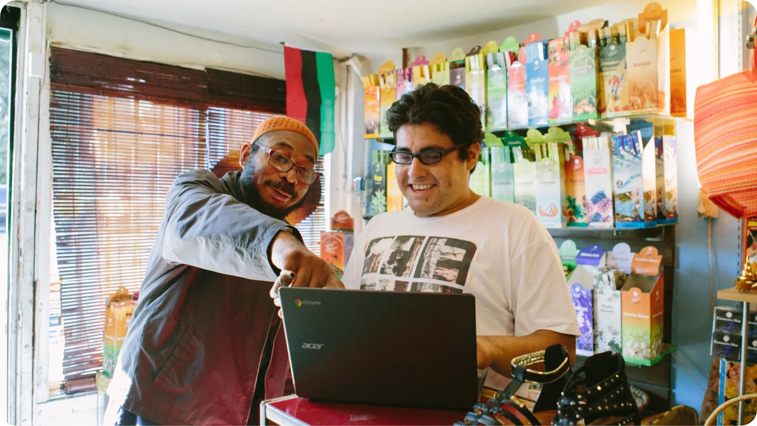 Two men smiling and looking at an Acer Chromebook laptop inside a retail shop or small business. One man, wearing an orange knit cap and glasses, points at the screen while the other man, wearing glasses and a graphic t-shirt, looks on. Shelves of packaged goods fill the background.