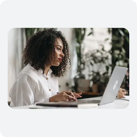 A woman with curly dark hair and a white button-down shirt looks intently at her laptop screen while typing at a white desk. In the blurred background, green houseplants are visible, creating a bright and modern remote work or office environment.