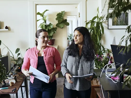 Two women walking side-by-side through a bright, modern office filled with green houseplants while holding documents and discussing work.